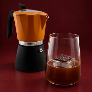 Brown cocktail with a large clear ice cube topped with a thin dark chocolate square, beside an orange moka pot on a burgundy background.