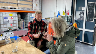Two people sitting at a table in a warehouse setting, clinking glasses.