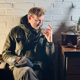 Hugh Anderson, in a green jacket sitting in a rustic setting with a white brick wall smelling a spirit prior to sampling it.