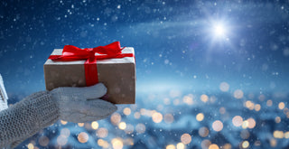 Hand wearing a gray glove holding a gift box with a red ribbon against a snowy night sky.