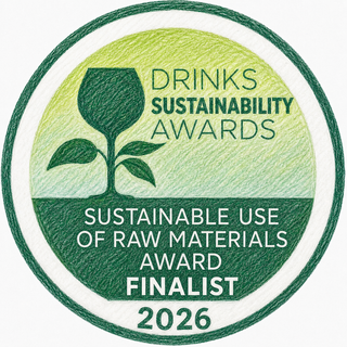 A circular award badge for the Drinks Sustainability Awards, recognizing the Sustainable Use of Raw Materials Award Finalist 2026. The badge features a green and yellow-green textured background with a dark green wine glass silhouette and a small plant sprout, surrounded by a dark green circular border on a white background.