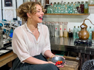 Woman sitting at a bar holding a bowl of fruit, with bottles and bar equipment in the background.
