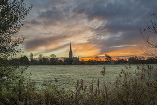 A scenic landscape photograph of a church spire on the horizon over a calm body of water, framed by reeds in the foreground and a dramatic sunset sky filled with orange, yellow, and gray clouds.