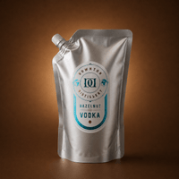 A silver stand-up pouch of Hazelnut Vodka, featuring a white and teal label, set against a warm brown gradient background.