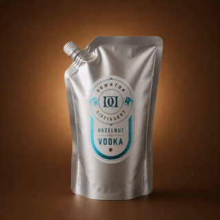 A silver stand-up pouch of Hazelnut Vodka, featuring a white and teal label, set against a warm brown gradient background.